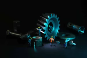 Des petits ouvriers portant des casques de sécurité travaillent sur des écrous, boulons et engrenages de grande taille, illustrant une méthode de maintenance industrielle appelée QQOCQCCP.