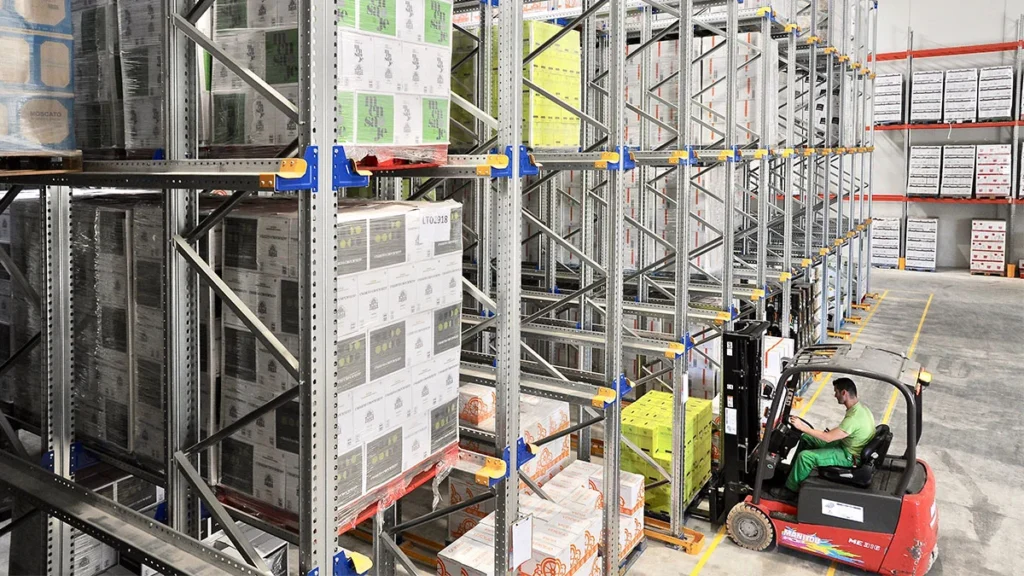 Un cariste conduit un chariot élévateur dans un entrepôt moderne, déplaçant des palettes de marchandises empilées sur de hauts rayonnages métalliques.