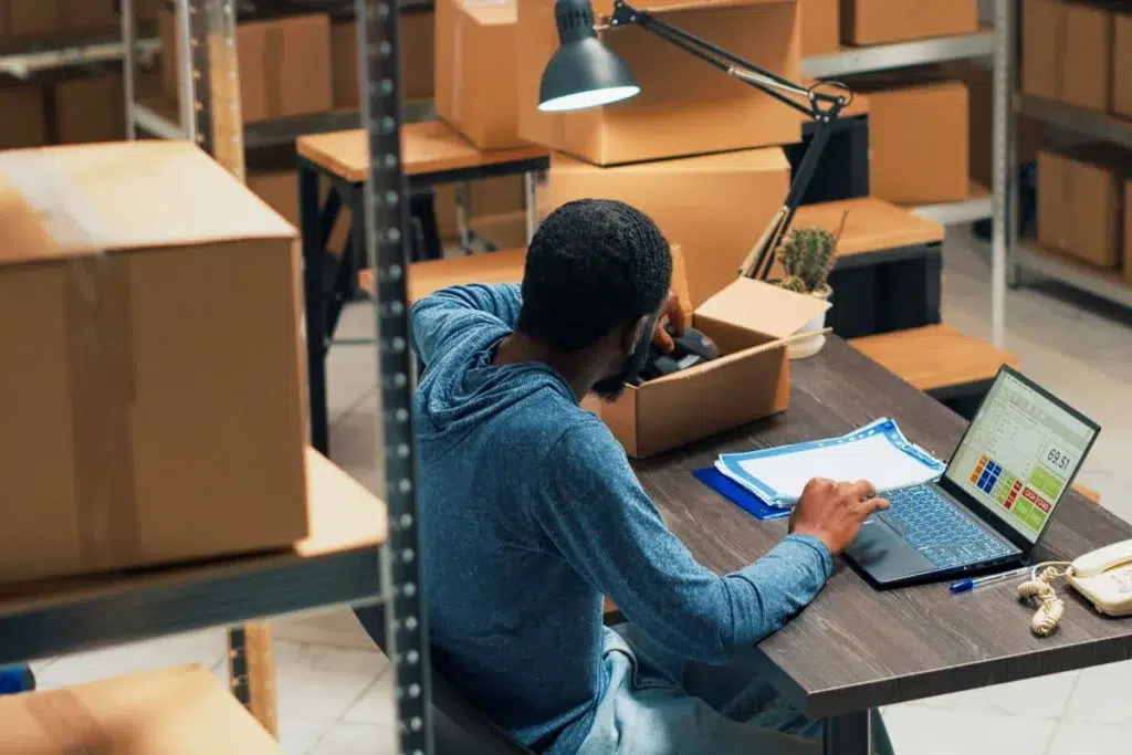 Homme de dos gérant des commandes sur ordinateur portable entouré de colis dans un petit entrepôt ou espace de stockage e-commerce