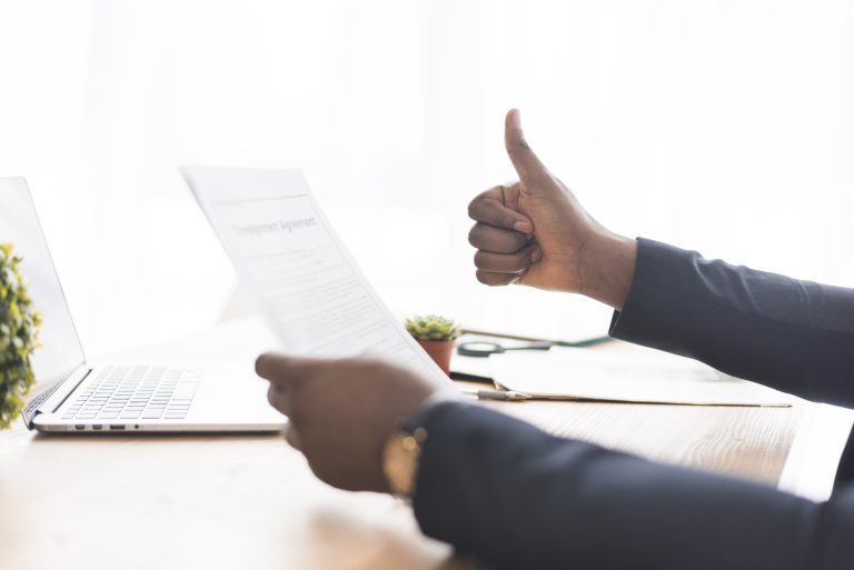 Professionnel tenant un document contractuel et faisant un pouce levé devant un ordinateur portable