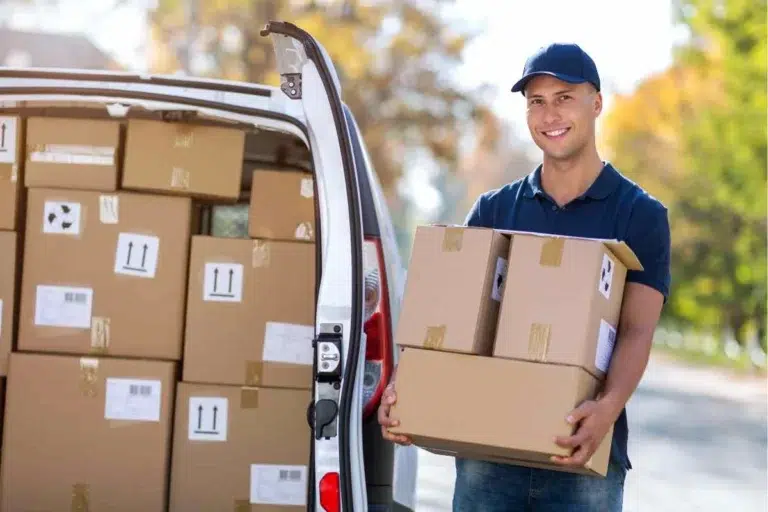 Livreur souriant en polo bleu et casquette tenant des colis devant un fourgon blanc chargé de cartons
