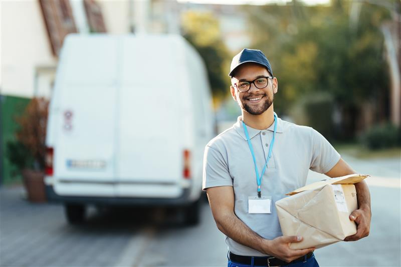 Livreur souriant portant des lunettes et une casquette, tenant un colis sous le bras devant son véhicule utilitaire blanc, en zone résidentielle