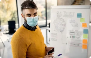 Homme en pull jaune portant un masque chirurgical tenant un smartphone devant un tableau blanc couvert de schémas et post-its colorés