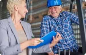 Manager en veste grise et opérateur avec casque bleu échangeant en souriant avec un clipboard dans un entrepôt logistique