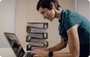 Femme souriante tapant sur un ordinateur portable avec une pile de classeurs colorés en arrière-plan symbolisant les documents papier à dématérialiser