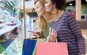 Deux femmes souriantes portant des sacs de shopping regardant une vitrine dans un centre commercial