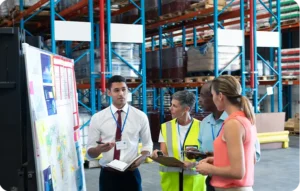 Équipe de quatre professionnels logistiques en réunion debout devant un tableau de bord avec cartes et graphiques dans un entrepôt