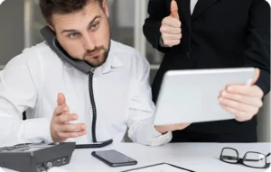 Conseiller client au téléphone fixe pendant qu'un collègue lui montre une tablette en faisant un pouce levé